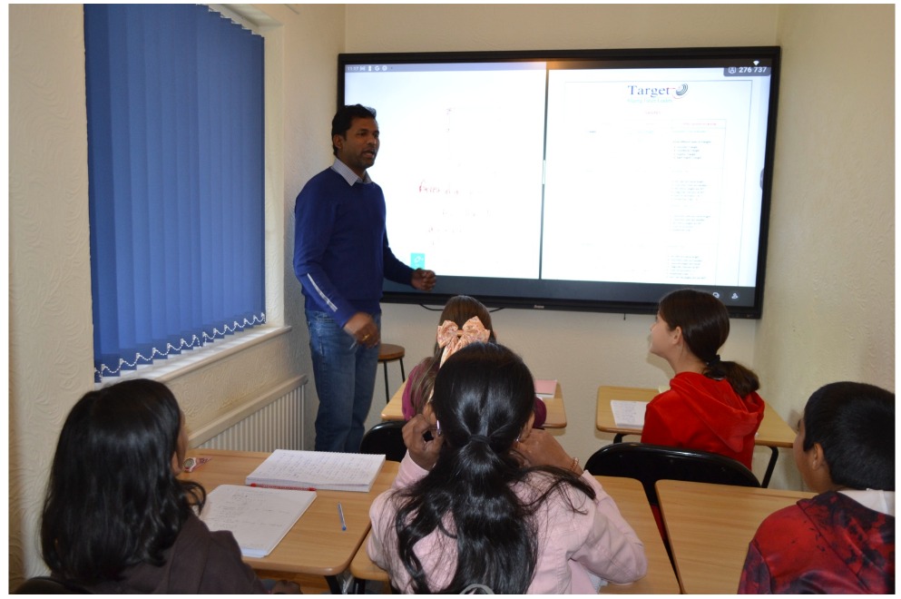 Male teacher in a blue sweater presents at the front of a classroom, with a large screen displaying a document.